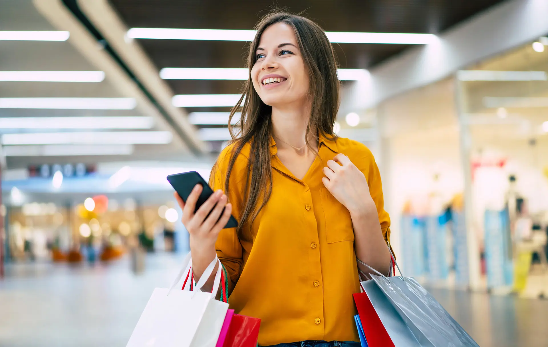 En glad kvinna shoppar i en galleria och håller sin telefon i handen.